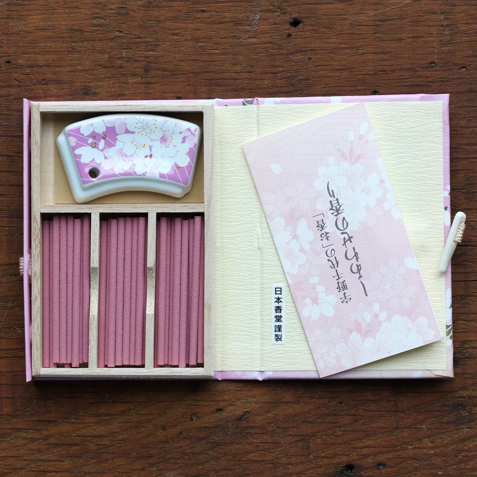 The Cherry Blossom Incense set includes pink incense sticks in a wooden gift box, a floral ceramic stand, and an open booklet featuring cherry blossom designs and Japanese text, all beautifully arranged on a wooden surface.