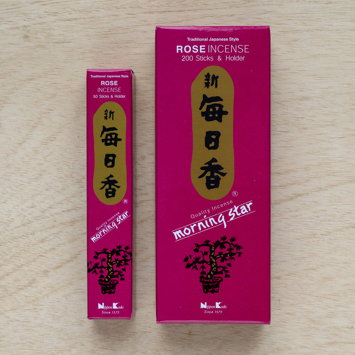 Two dark pink boxes of Morning Star Rose Incense with gold and white text rest on wood. The larger box, featuring Japanese characters, holds 200 sticks and a holder; the smaller contains 50. Both showcase a pure floral essence blend.