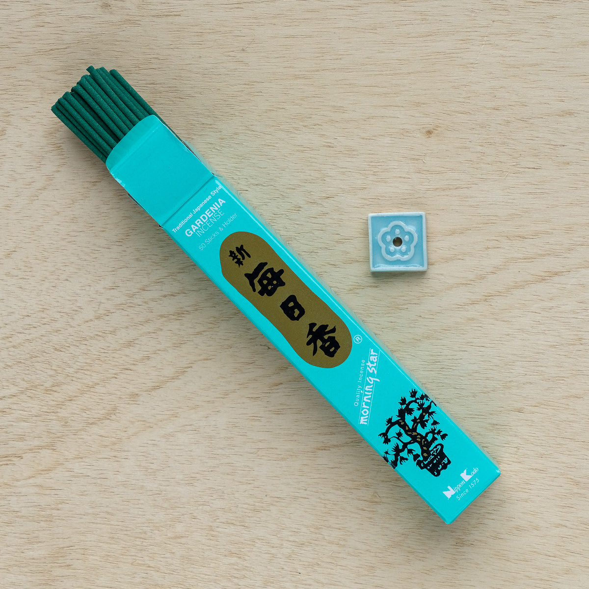 A turquoise box of Morning Star Gardenia Incense sits on wood, revealing aromatic sticks inside. Accompanied by a small square holder with a floral design, its perfect for a pure floral experience.