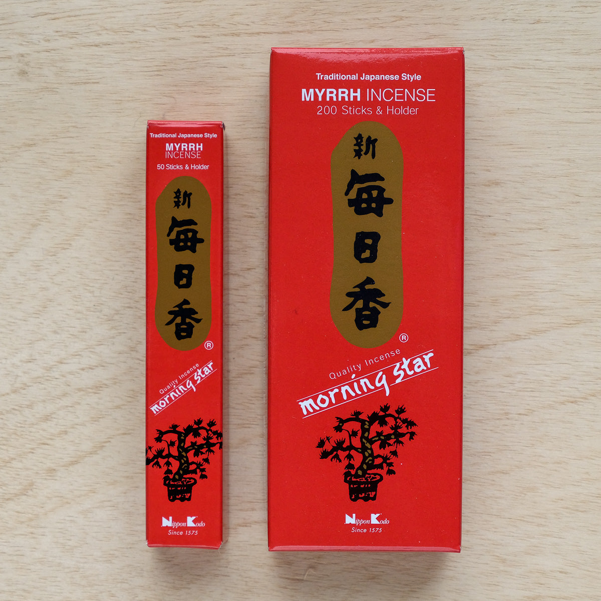 On a wooden surface lie two boxes of Morning Star Myrrh Incense, each releasing a rich Japanese myrrh scent. The larger box contains 200 sticks with an elegant holder, while the smaller has 50. Both boxes are striking red with Japanese text and floral designs.