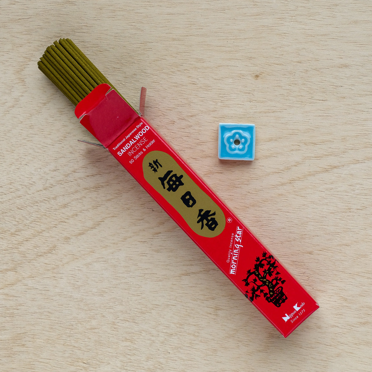 A red box of Morning Star Sandalwood Incense lies on a wooden surface, with one stick partly removed, releasing a calming scent. Nearby, a small blue tile featuring a white floral design adds an artistic touch.