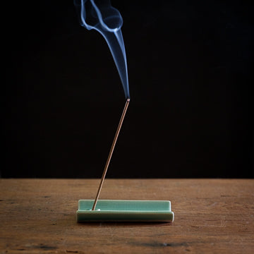 Incense stick with smoke on a green tray against a black background