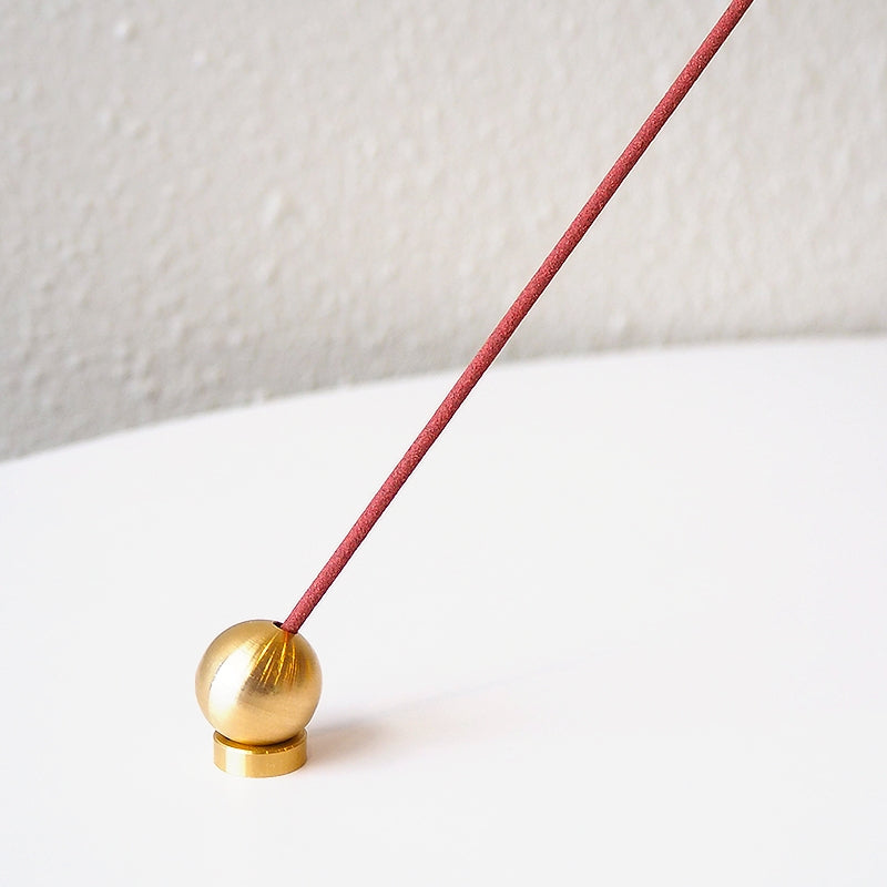 A red incense stick is elegantly angled into a small Sphere Incense Holder made of brass, resting on a white surface against a light gray background.