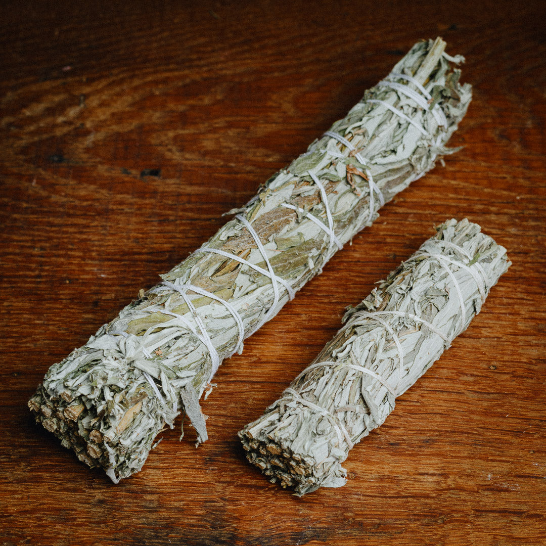 Two bundled Black Sage (Mugwort) smudge sticks, differing in length, are wrapped in twine on a wooden surface. The tightly bound dried sage leaves, ideal for cleansing rituals, add a rich aroma and depth to the ceremony.
