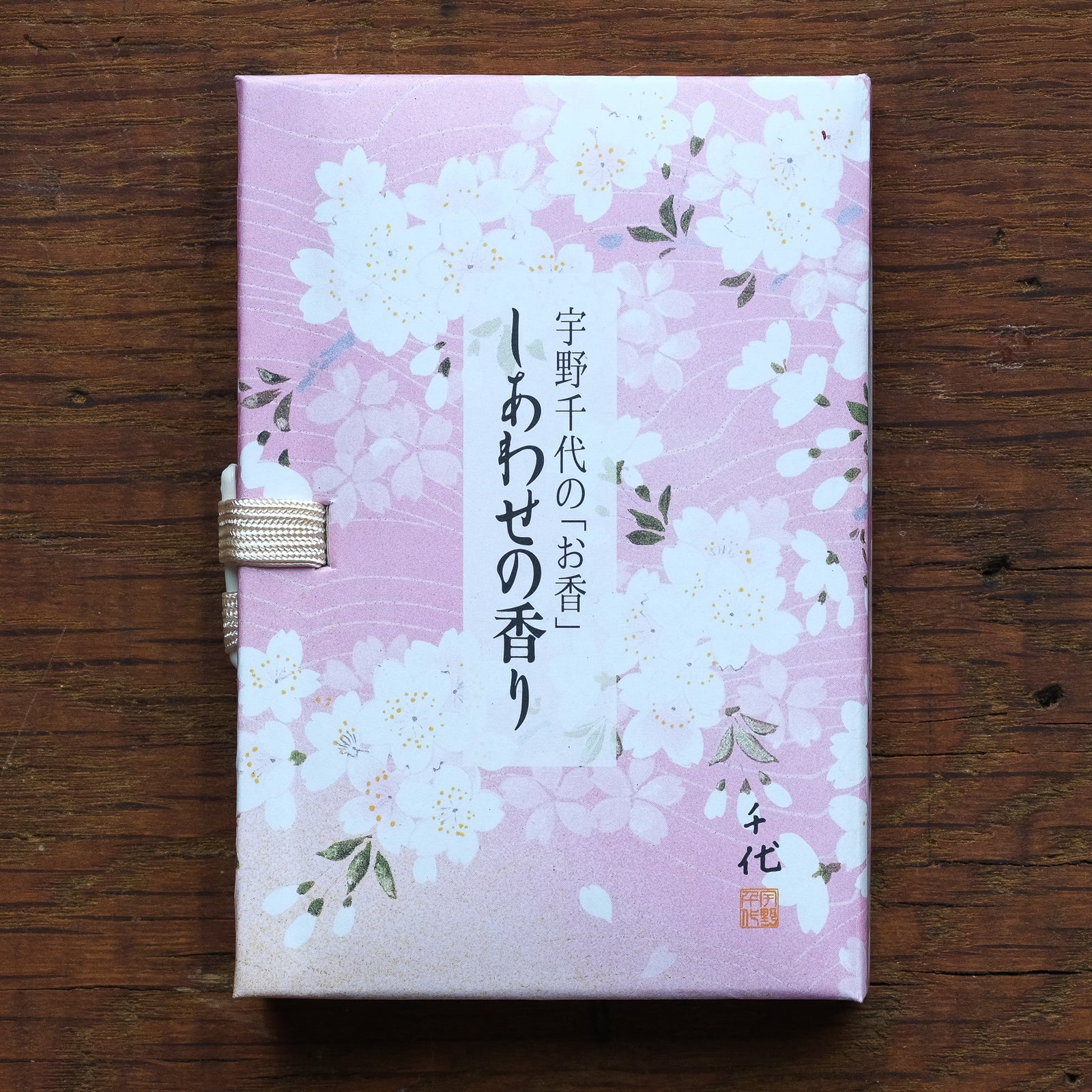 On a wooden surface rests the Cherry Blossom Incense box, featuring a pink cherry blossom design and Japanese text. Next to it is a ceramic stand for Japanese-style incense, adorned with serene white and pink floral patterns.
