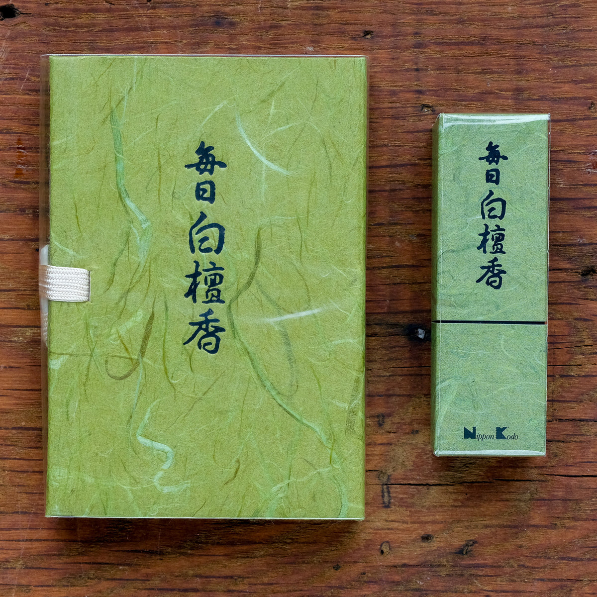 Two green, textured packages with Japanese writing rest on a wooden surface. The larger Mainichi Byakudan Premium Sandalwood Japanese Incense package, perfect for meditation, features a white ribbon. The smaller incense box stands upright beside it, and the small logo reads nYpon Kodo.
