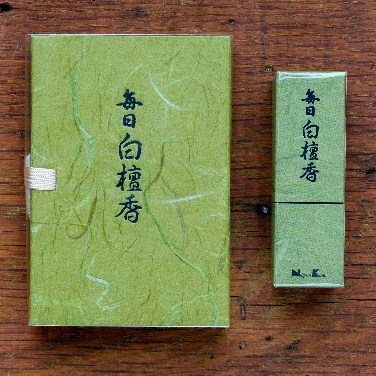 Two green, textured packages with Japanese writing rest on a wooden surface. The larger Mainichi Byakudan Premium Sandalwood Japanese Incense package, perfect for meditation, features a white ribbon. The smaller incense box stands upright beside it, and the small logo reads nYpon Kodo.