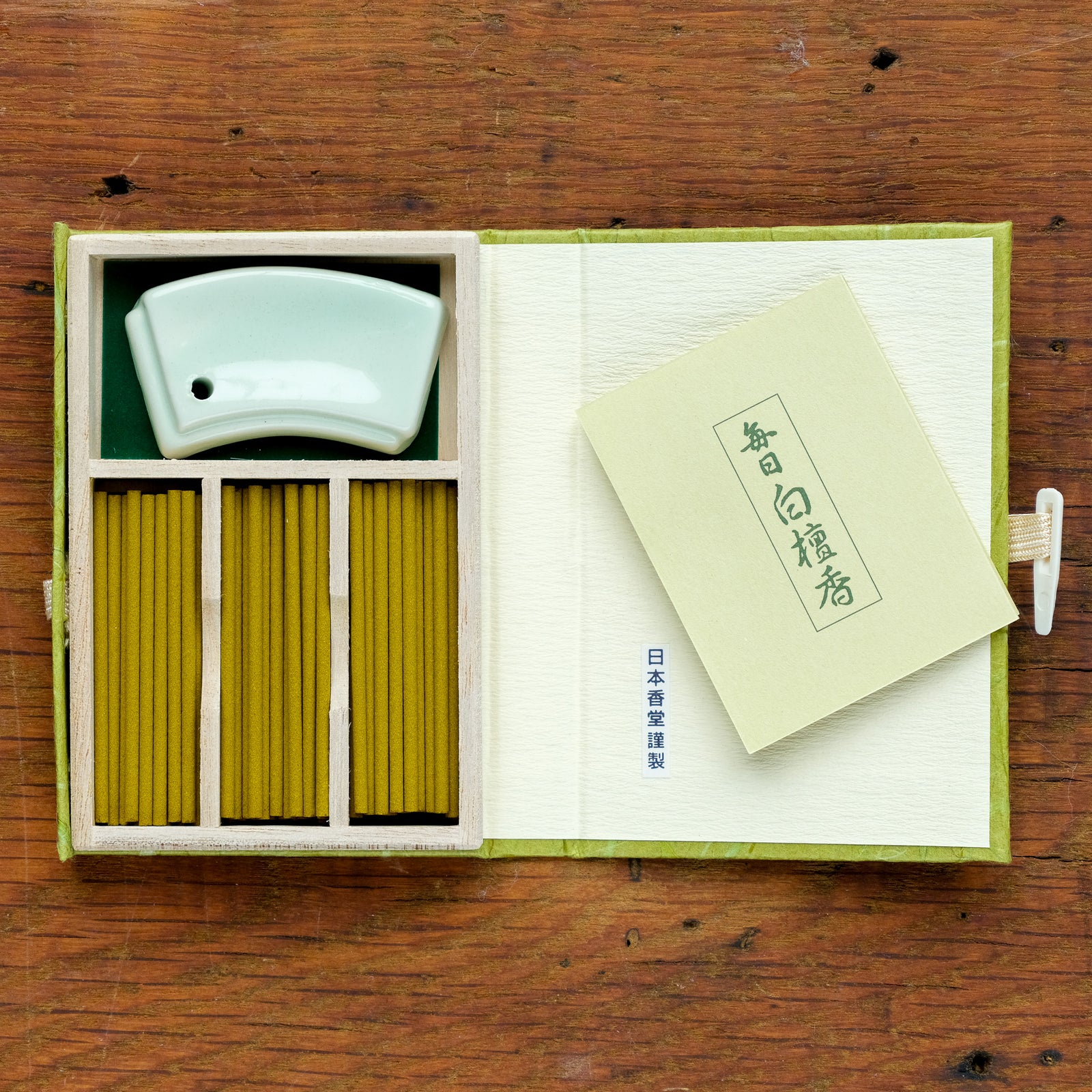 The Mainichi Byakudan Premium Sandalwood Japanese Incense set includes green sandalwood sticks, a white ceramic holder, and a booklet with Japanese text, all in an open box on wood. Ideal for meditation, the box features a green cover and a white tie closure.