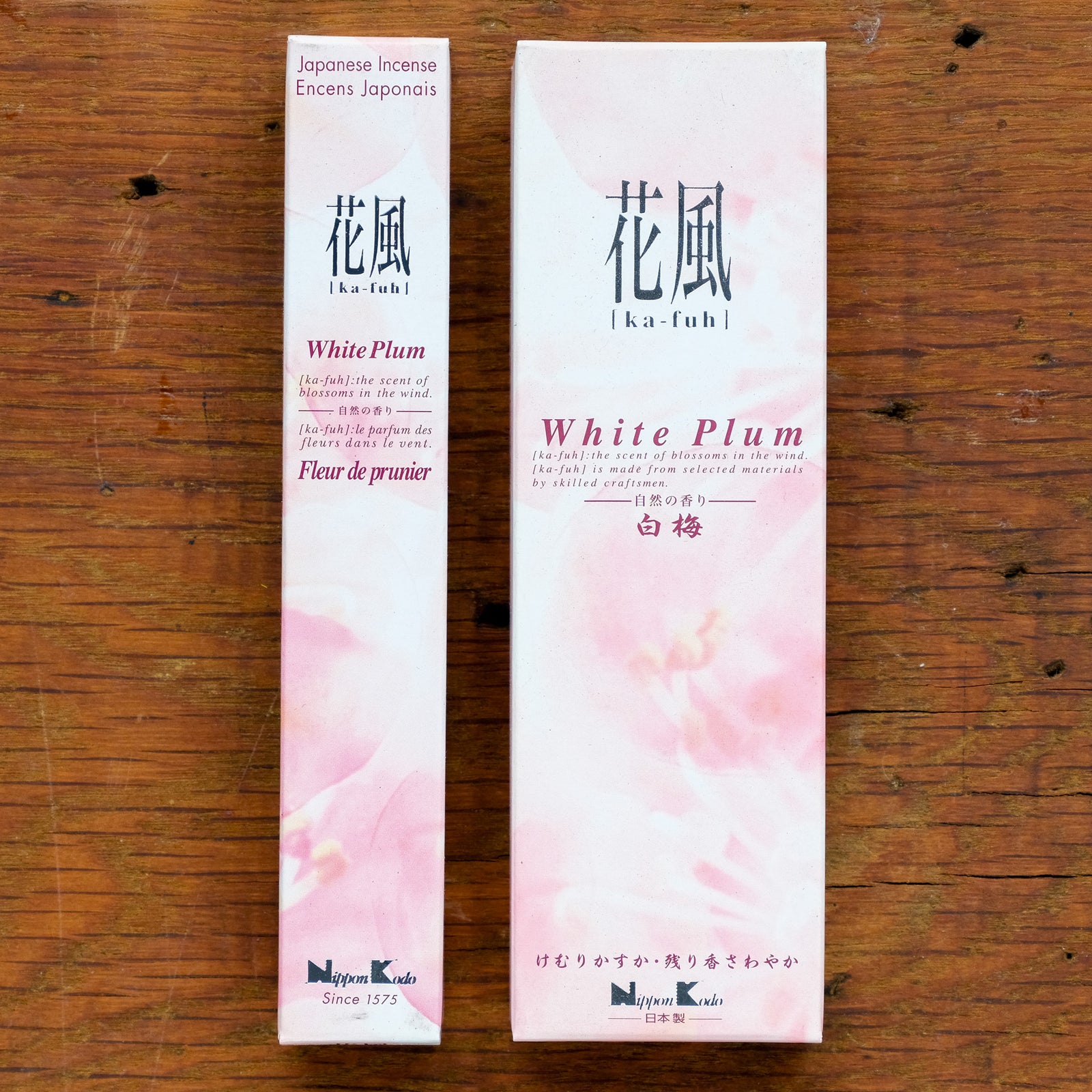 Two Ka-Fuh White Plum Incense boxes are elegantly displayed on wood. The taller box has pink and white floral designs with Japanese and English text. The shorter one matches perfectly, capturing the pure white plum fragrance.