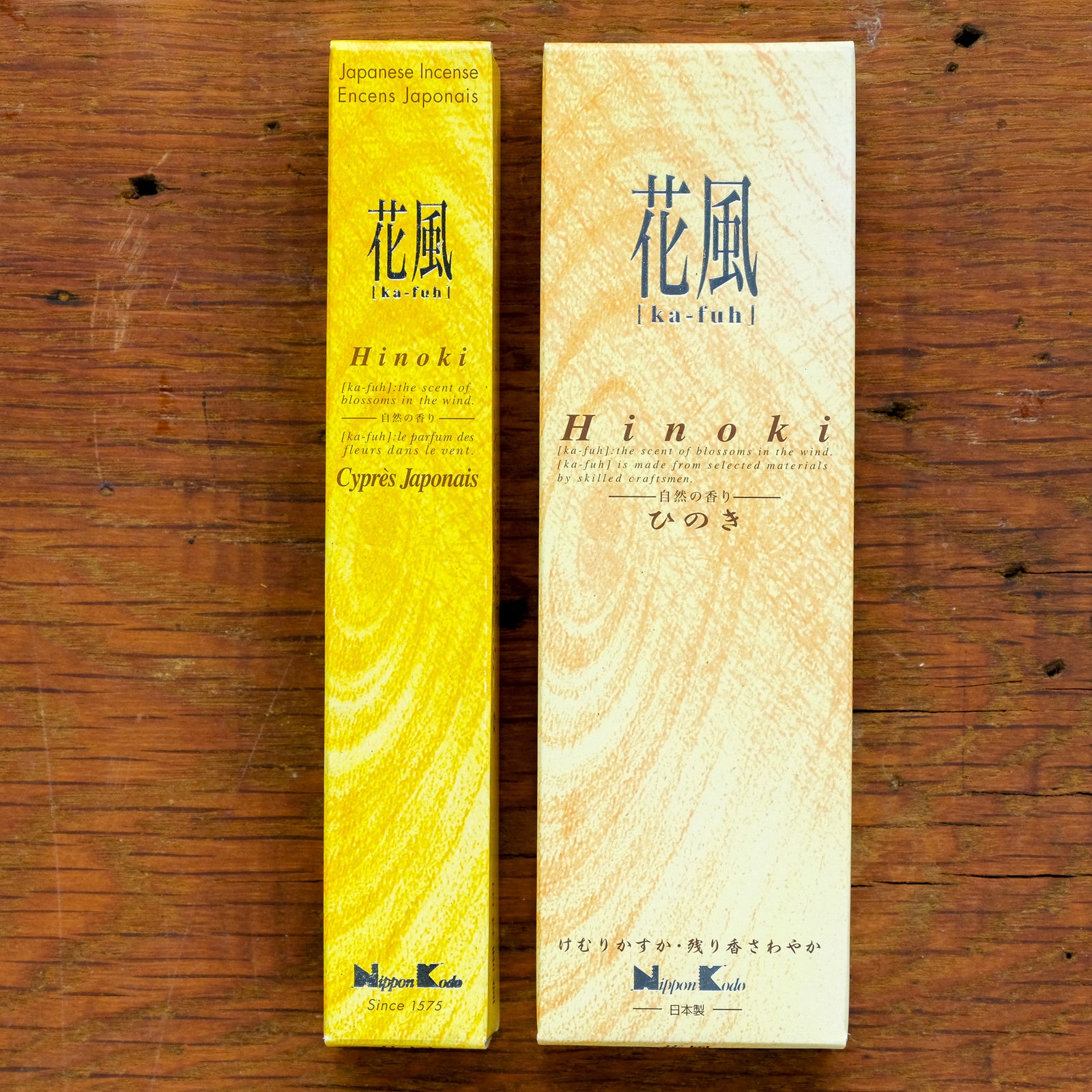 Two boxes of Ka-Fuh Hinoki (Japanese Cypress) Incense, in stylish typography, exude a pure scent. The yellow and cream boxes on a wooden surface evoke the hinoki aroma, with Japanese text about Cypress Japonais adding authenticity.