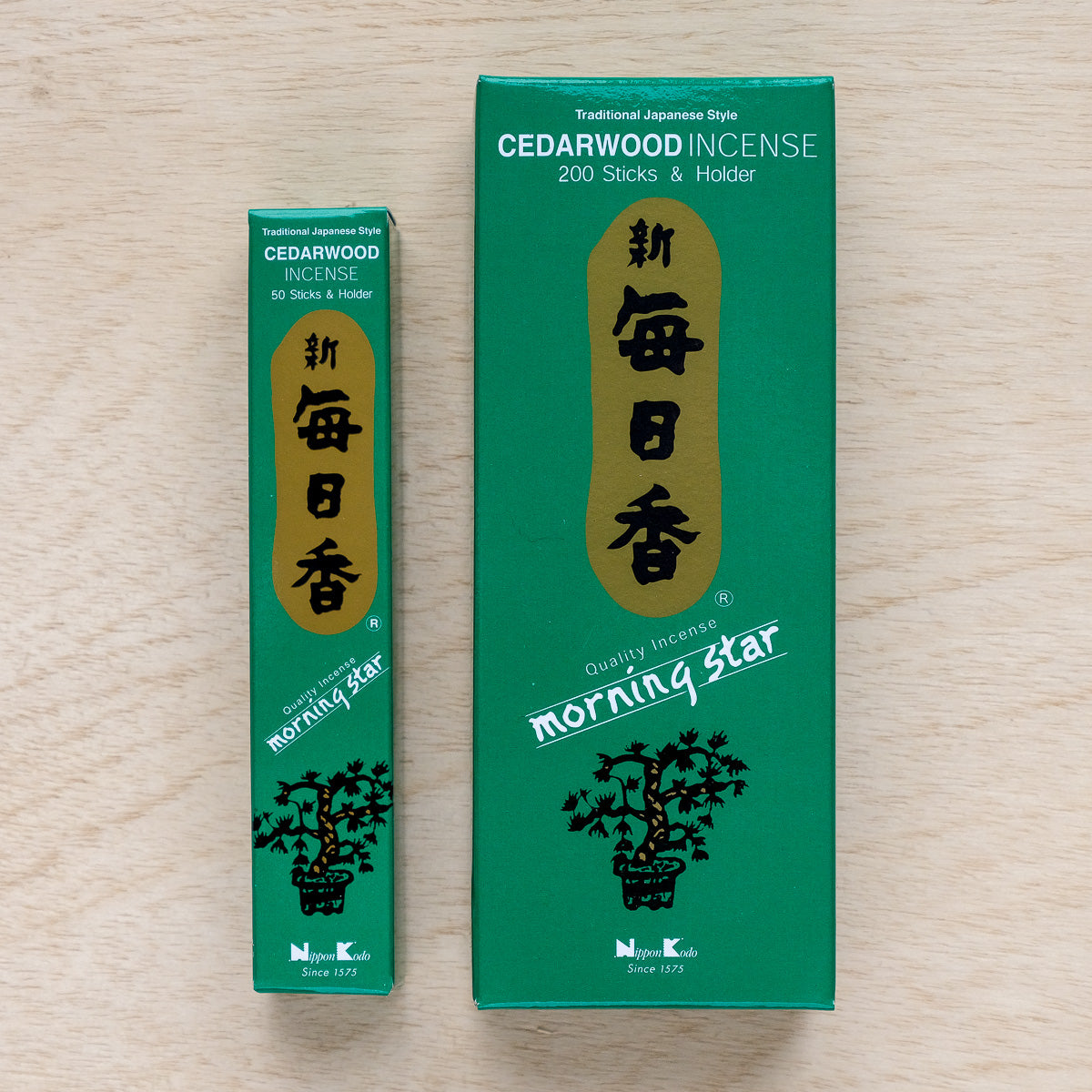 Two green boxes of Morning Star Cedarwood Incense, with gold and white text, rest on a wooden surface. The larger box contains 200 sticks and a holder, the smaller has 50. Both feature Japanese-style design with traditional characters, ideal for meditation rituals.
