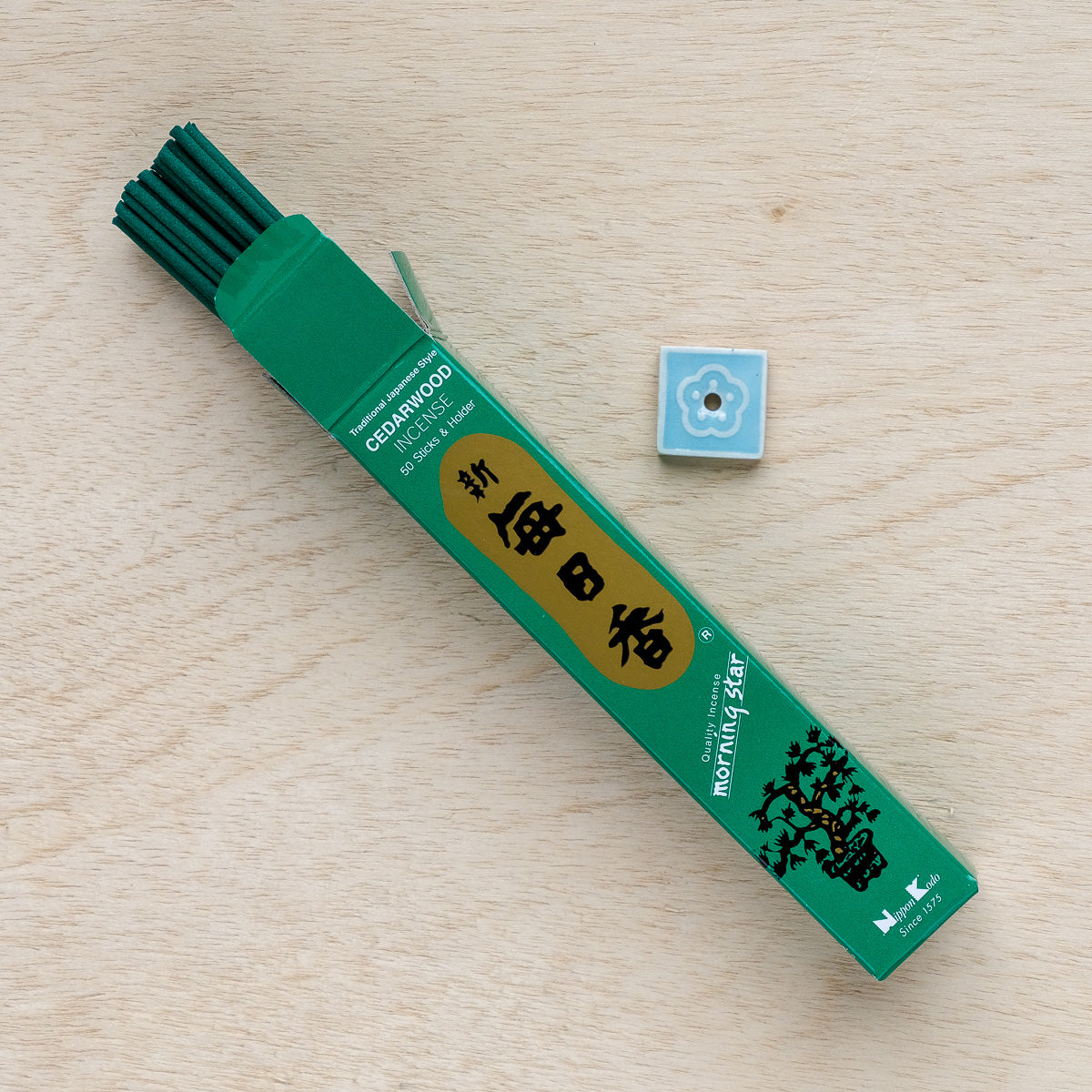An open green box of Morning Star Cedarwood Incense, celebrated for its Japanese style, rests on a wooden surface. Visible cedarwood sticks are beside a small blue floral-patterned ceramic holder ideal for meditation.