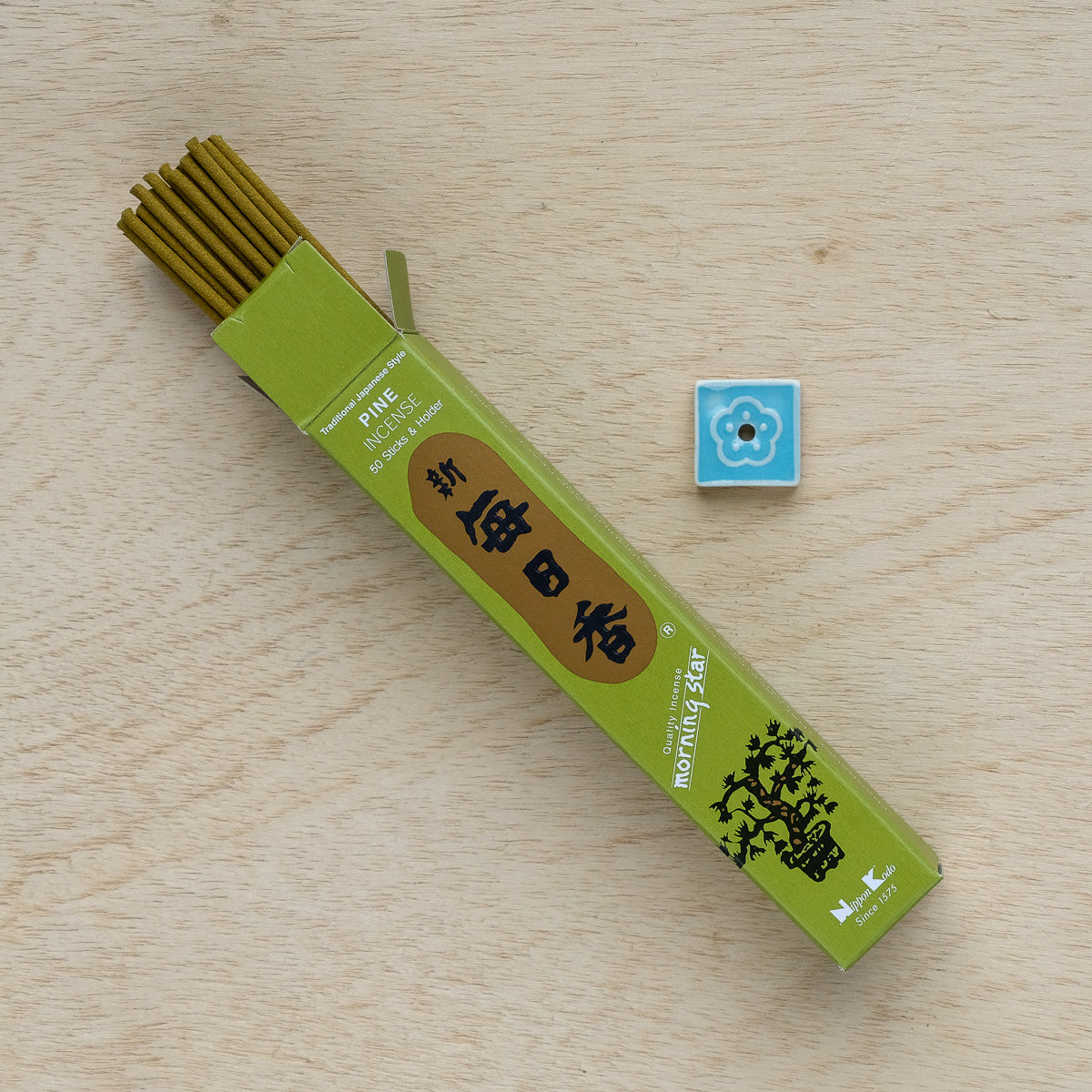 A box of Morning Star Pine Incense lies open on a wooden surface, releasing a calming pine aroma. Several sticks are partially visible next to a square, blue ceramic tile with a white flower design—creating an ideal setting for serene meditation moments.
