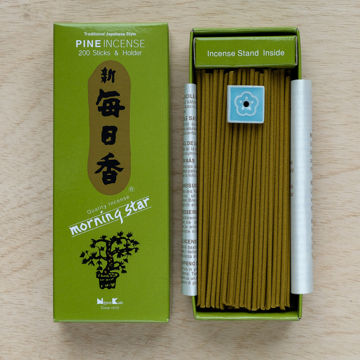 Place the open green box of Morning Star Pine Incense on a wooden surface. The package offers 200 pine sticks and includes a small, blue, flower-shaped holder. Traditional Japanese style packaging enhances meditation moments beautifully.
