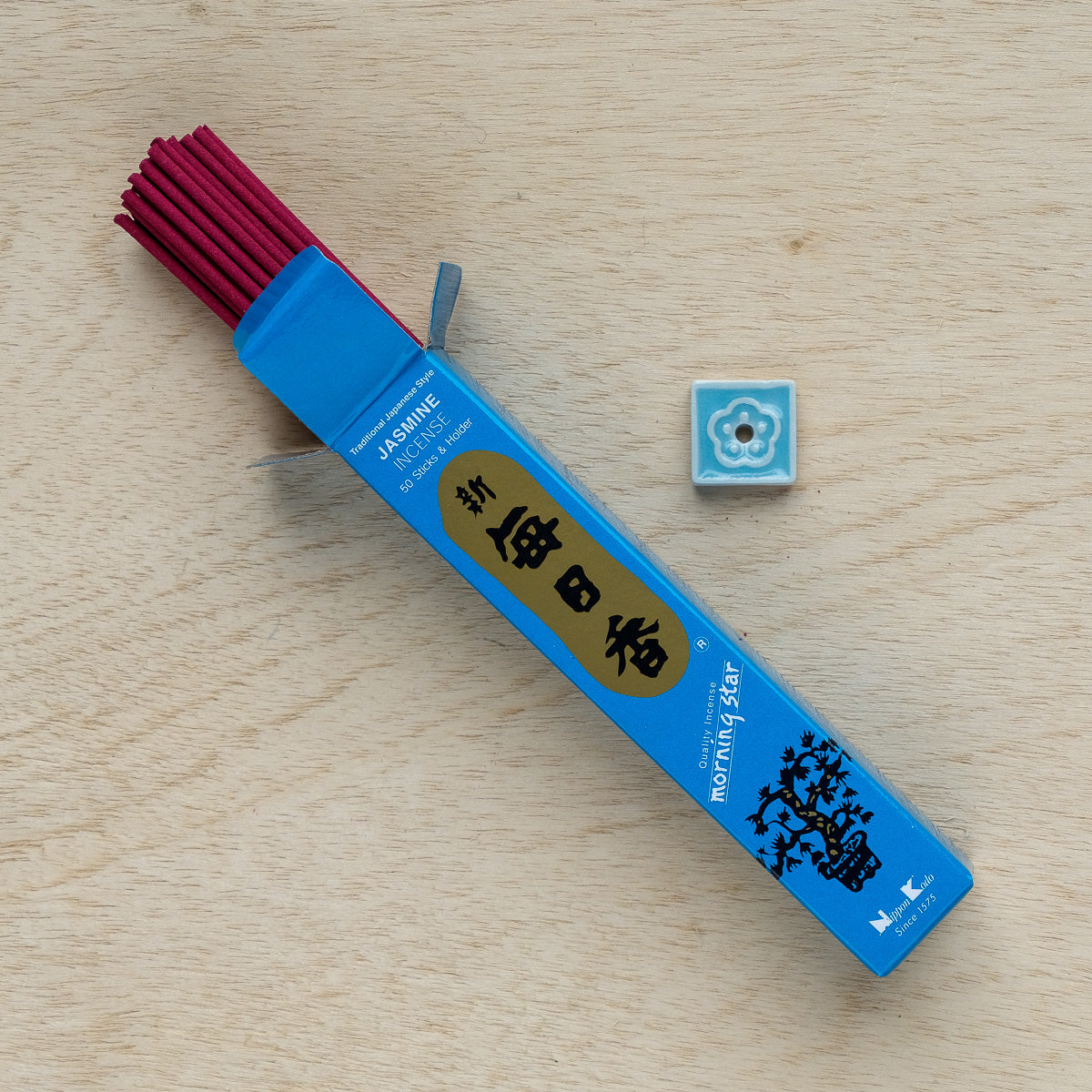An open box of Morning Star Jasmine Incense displays several red sticks. A blue ceramic holder with a white floral design sits next to it on light wood, perfect for enhancing meditation with its soothing aroma.