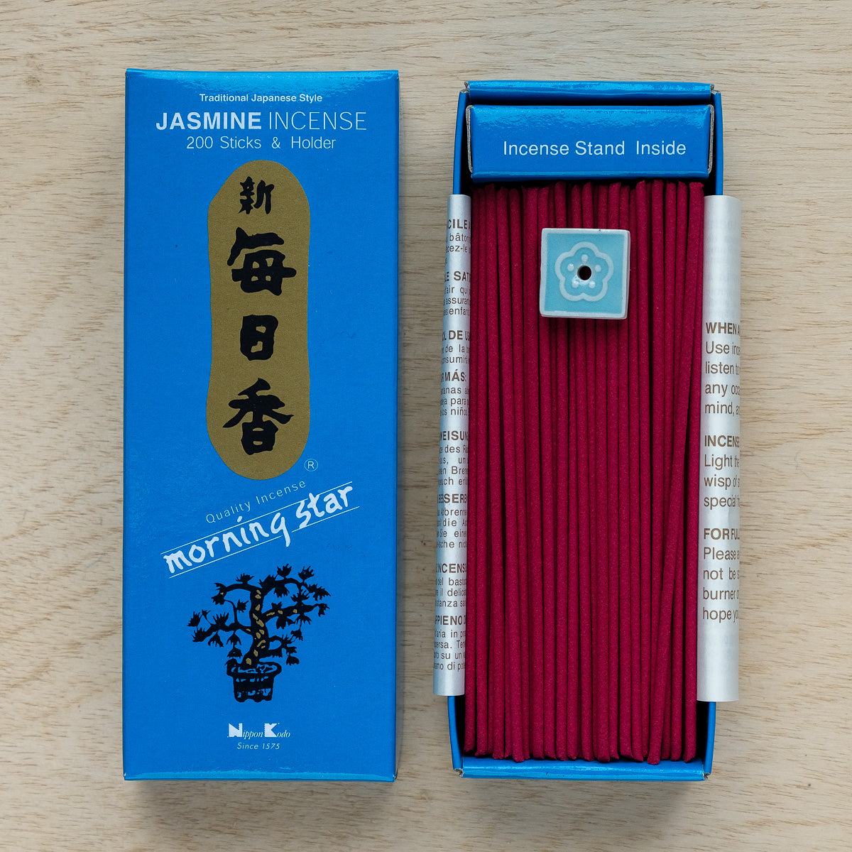 An open blue box of Morning Star Jasmine Incense rests on wood, showcasing red sticks and a delicate ceramic holder. Perfect for meditation, the Japanese packaging is adorned with text and a tree image.