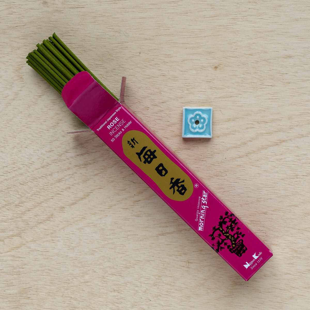 An open box of Morning Star Rose Incense sits on a wooden surface, with some sticks partially visible next to a small decorative blue tile featuring a delicate white floral pattern.