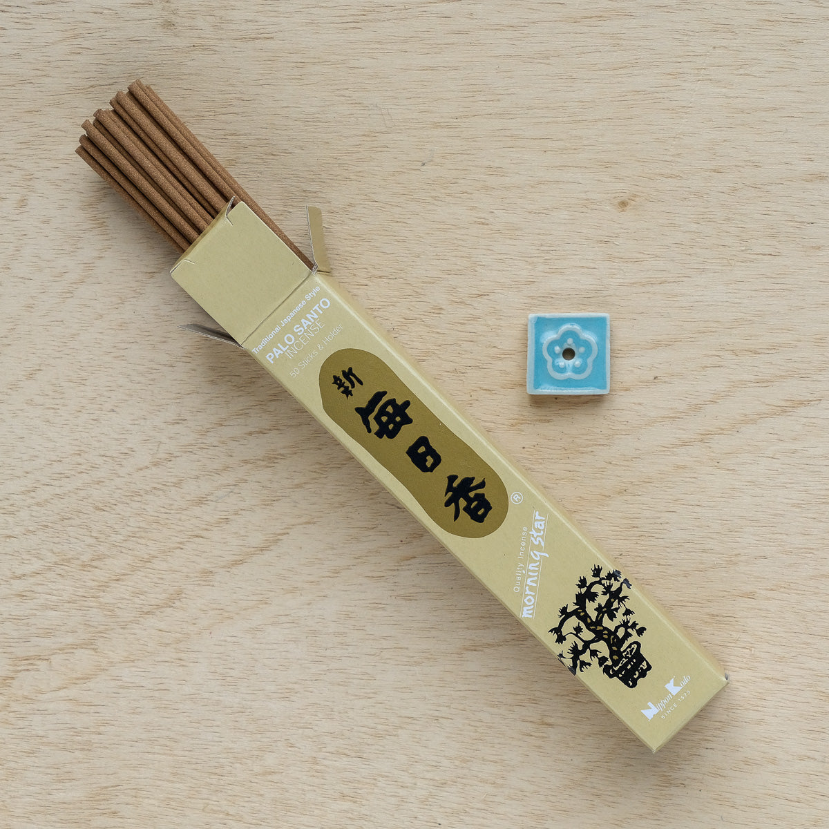 An open box of Morning Star Palo Santo Incense sits on a wooden surface next to a blue floral-patterned Japanese holder. The packaging displays text and a floral design, suggesting the rich frankincense scent inside.