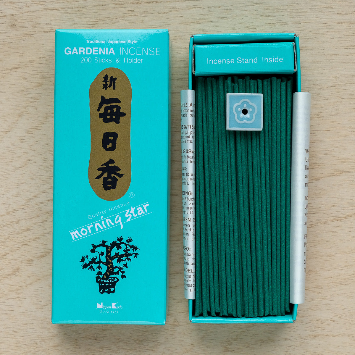 The open box of Morning Star Gardenia Incense rests on wood, revealing 200 green sticks and a small holder. With pure floral essences, the turquoise packaging showcases gold and black Japanese-style designs, embodying traditional Japanese incense.