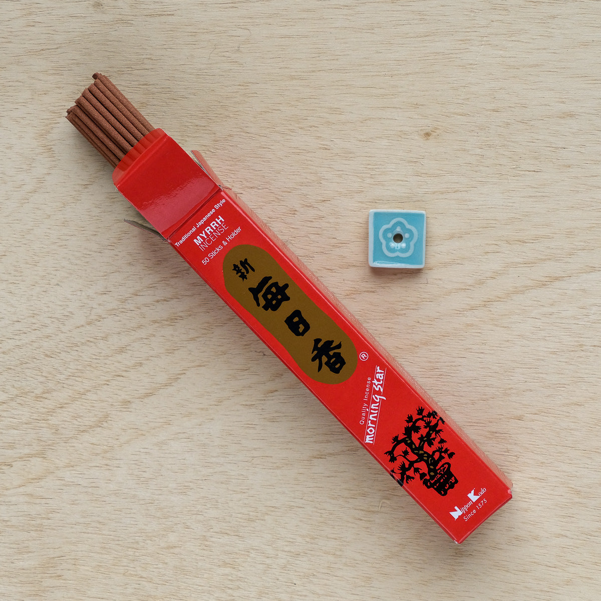 An open red box of Morning Star Myrrh Incense lies on wood, revealing several sticks. Beside it is a small square ceramic holder with a blue floral design, ready to release the soothing myrrh fragrance into the air.