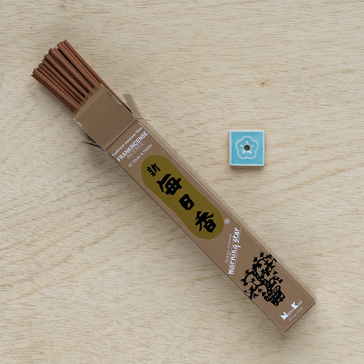 A box of Morning Star Frankincense Incense with a floral design and Japanese text lies open on wood. Beside it sits a small square blue tile featuring a white flower design, epitomizing tranquility.