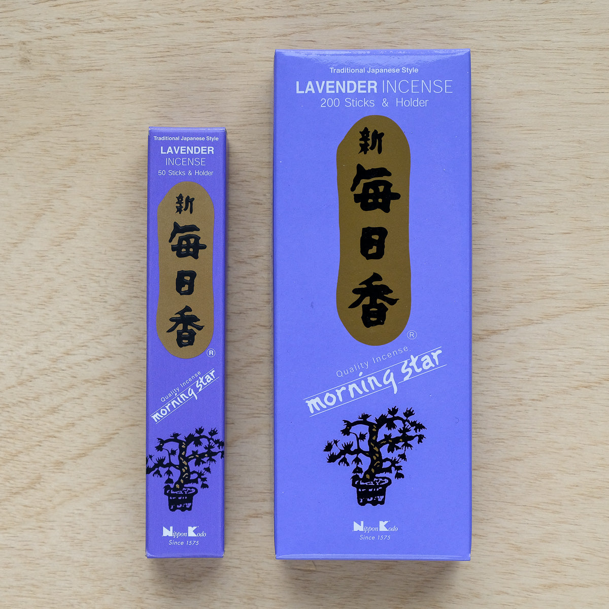 Two boxes of Morning Star Lavender Incense, infused with aromatic woods, rest on a wooden surface. The smaller box contains 50 sticks, while the larger contains 200 sticks and a holder. Both feature a Japanese style with purple hues and gold text.