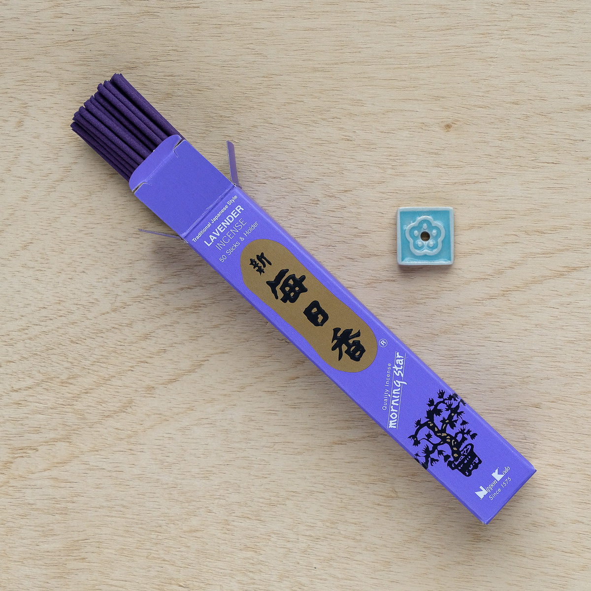 Purple incense sticks rest near an open Morning Star Lavender Incense box on a wooden surface. A small, decorative ceramic tile with a blue floral design nearby adds to the serene elegance of Japanese-style incense.