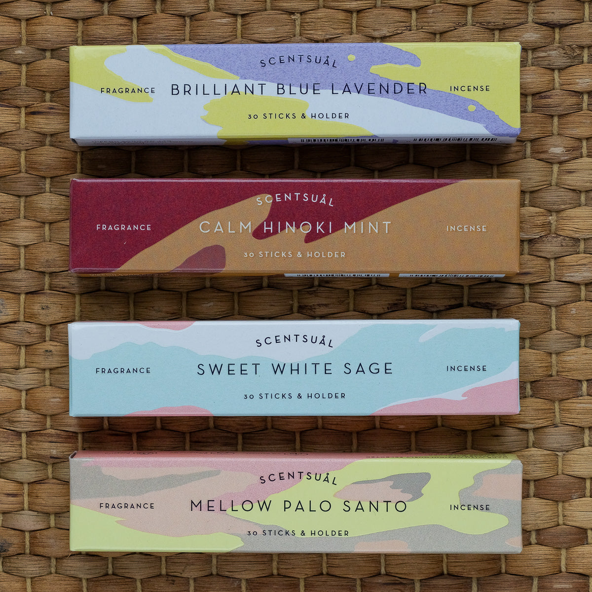 Four boxes of Scentsual Japanese Incense are stacked, labeled from top to bottom as Brilliant Blue Lavender, Calm Hinoki Mint, Sweet White Sage, and Mellow Palo Santo. Each box offers a colorful design with 30 sticks and a holder for a clean fragrance experience.
