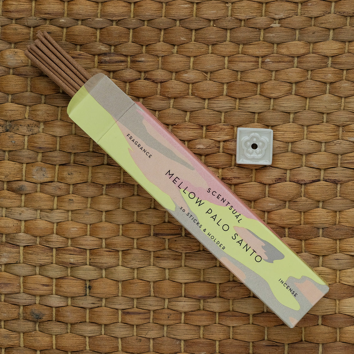 A box of Scentsual Japanese Incense labeled Mellow Palo Santo lies open on a woven surface, with incense sticks partially out. A thin square ceramic holder with a small hole sits nearby, emphasizing the natural fragrance and minimal packaging design of this incense.
