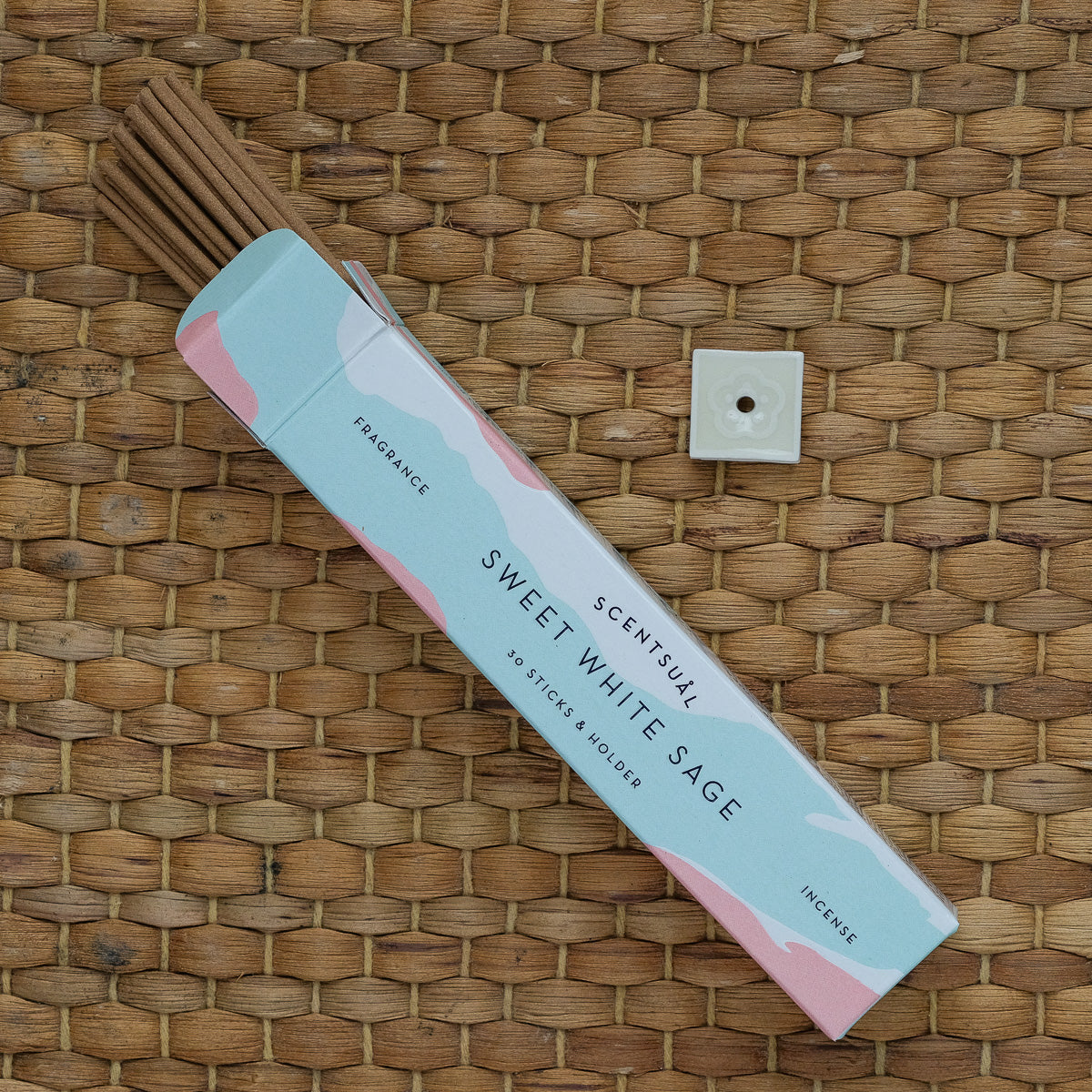 The Scentsual Japanese Incense box is open on a woven surface, releasing a clean fragrance. Several sticks are partially visible with a small square incense holder. The minimal packaging uses pastel colors and prominently displays FRAGRANCE and INCENSE.