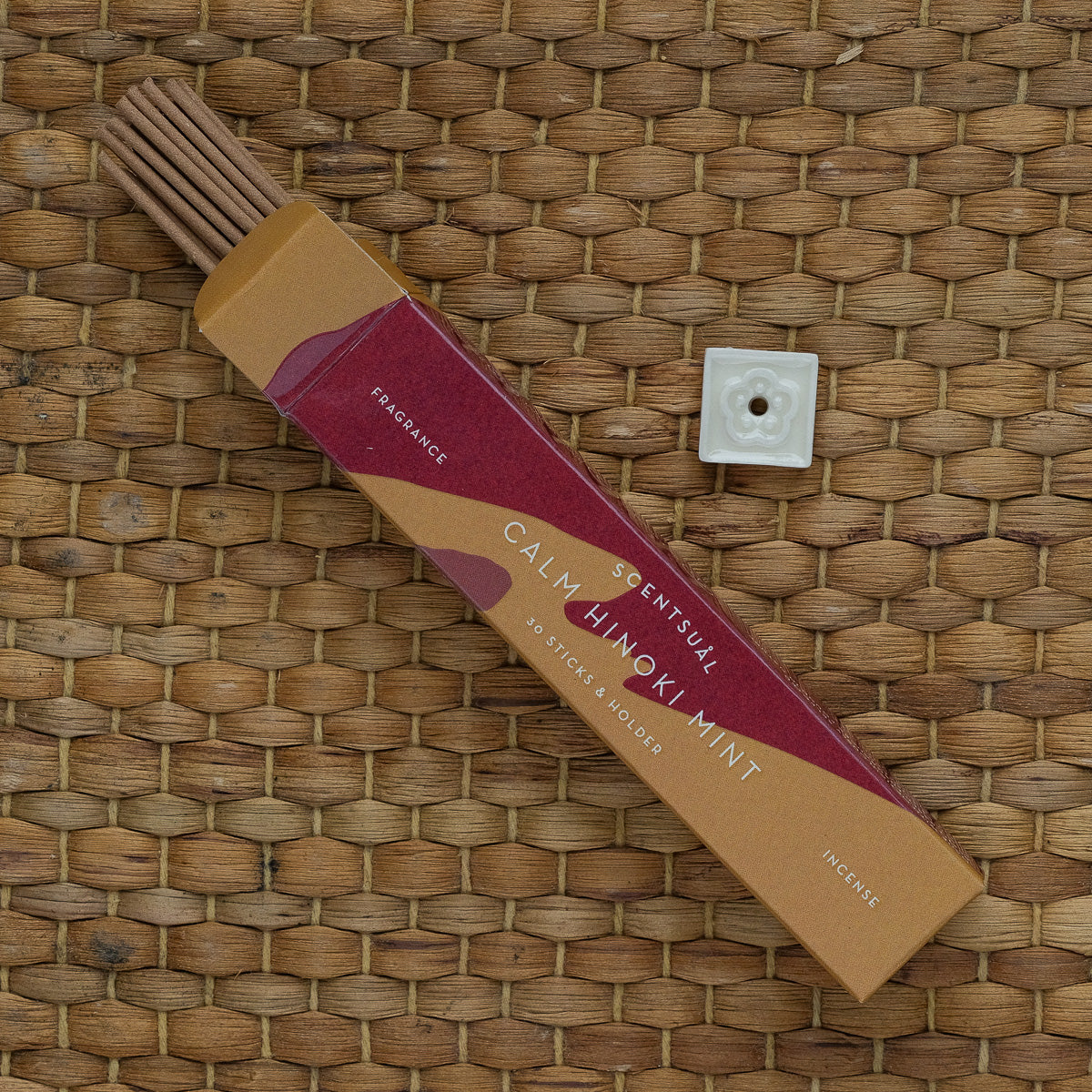 A box labeled Scentsual Japanese Incense in Calm Hinoki Mint scent is open on a woven surface. Nearby, theres a small square incense holder. The red and brown minimalist packaging highlights the clean fragrance and quantity of the natural incense sticks.