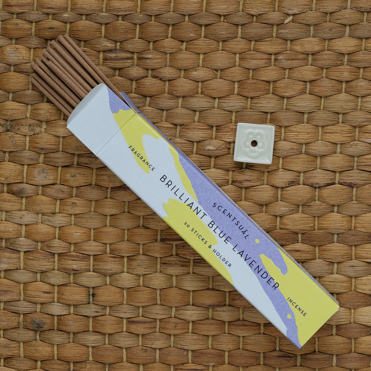 A box of Scentsual Japanese Incense with its clean fragrance is open on a woven surface, showing several sticks. A small white ceramic holder is beside it. The box has a yellow and purple design featuring text about its natural scent.