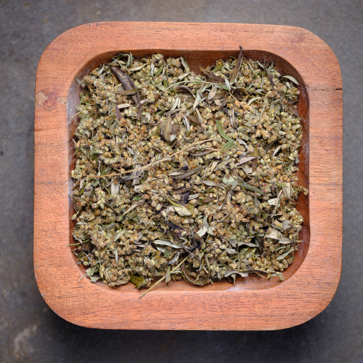 A square wooden bowl filled with Black Sage (Mugwort) Loose sits on a textured gray surface. The blend of small leaves and plant pieces creates a natural, earthy look.