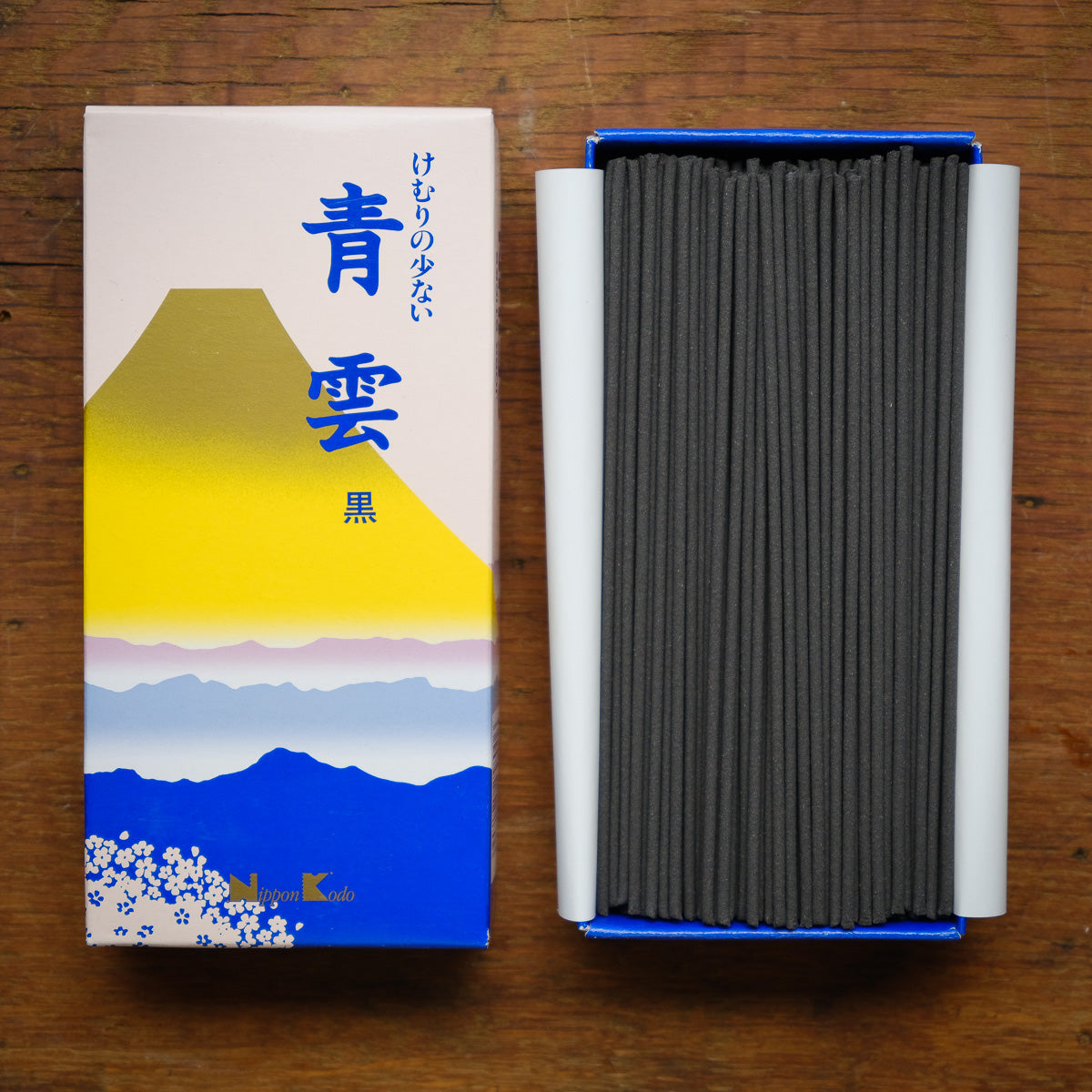 A box of Seiun Chrysanthemum incense features a yellow and blue mountain design, with its open side revealing neatly arranged black Nippon Kodo sticks on a wooden surface, offering a fragrant, low-smoke experience.