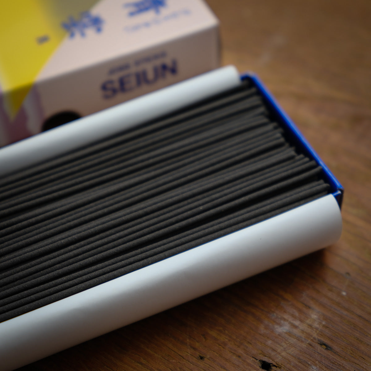 A close-up of Seiun Chrysanthemum Incense, known for its low-smoke formula, shows sticks in a white holder with Nippon Kodo packaging in the background. The box displays blue text and yellow-purple design on a brown wooden surface.