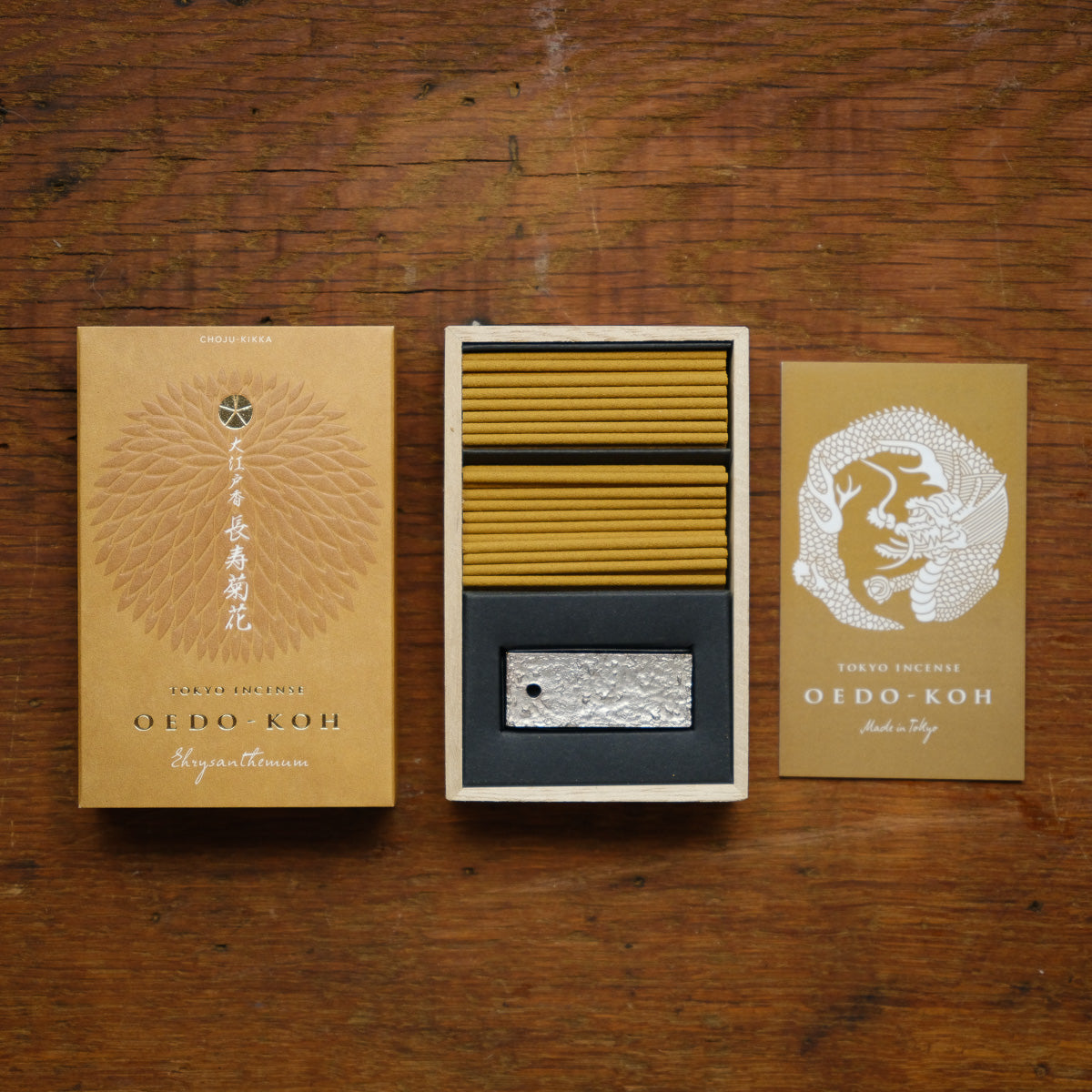 A box of Oedo-Koh Chrysanthemum Incense and a detailed metallic incense holder sit on a wooden surface. The packaging features elegant gold and brown designs, reminiscent of Ukiyo-e paintings, with Japanese script and a geometric pattern reflecting traditional Japanese style.