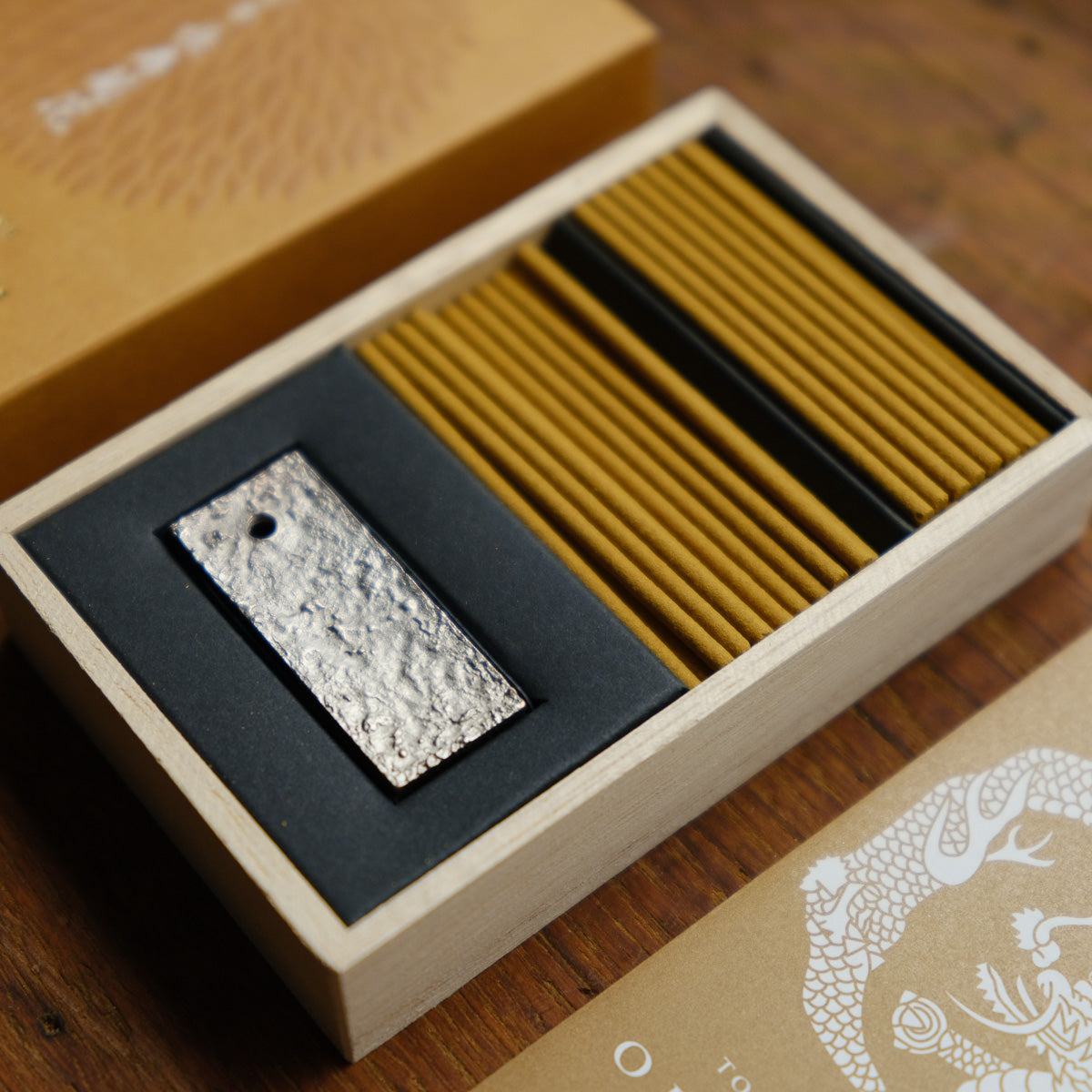 An Oedo-Koh Chrysanthemum Incense box with a black insert lies beside a textured metallic holder, evoking Ukiyo-e art. Both rest on wood, and the packaging showcases a dragon entwined with chrysanthemum motifs.