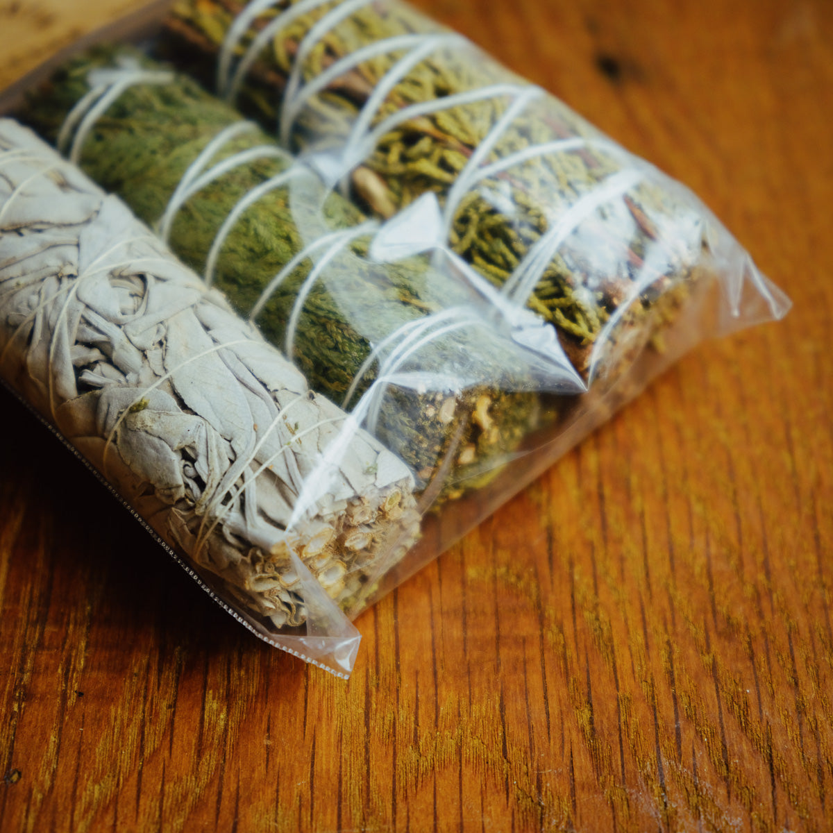A transparent plastic bag on a wooden surface holds the White Sage, Cedar, Juniper Smudge Incense Set. The herbs, bound with twine, showcase textures and colors ranging from silvery-green to dark green and brown.