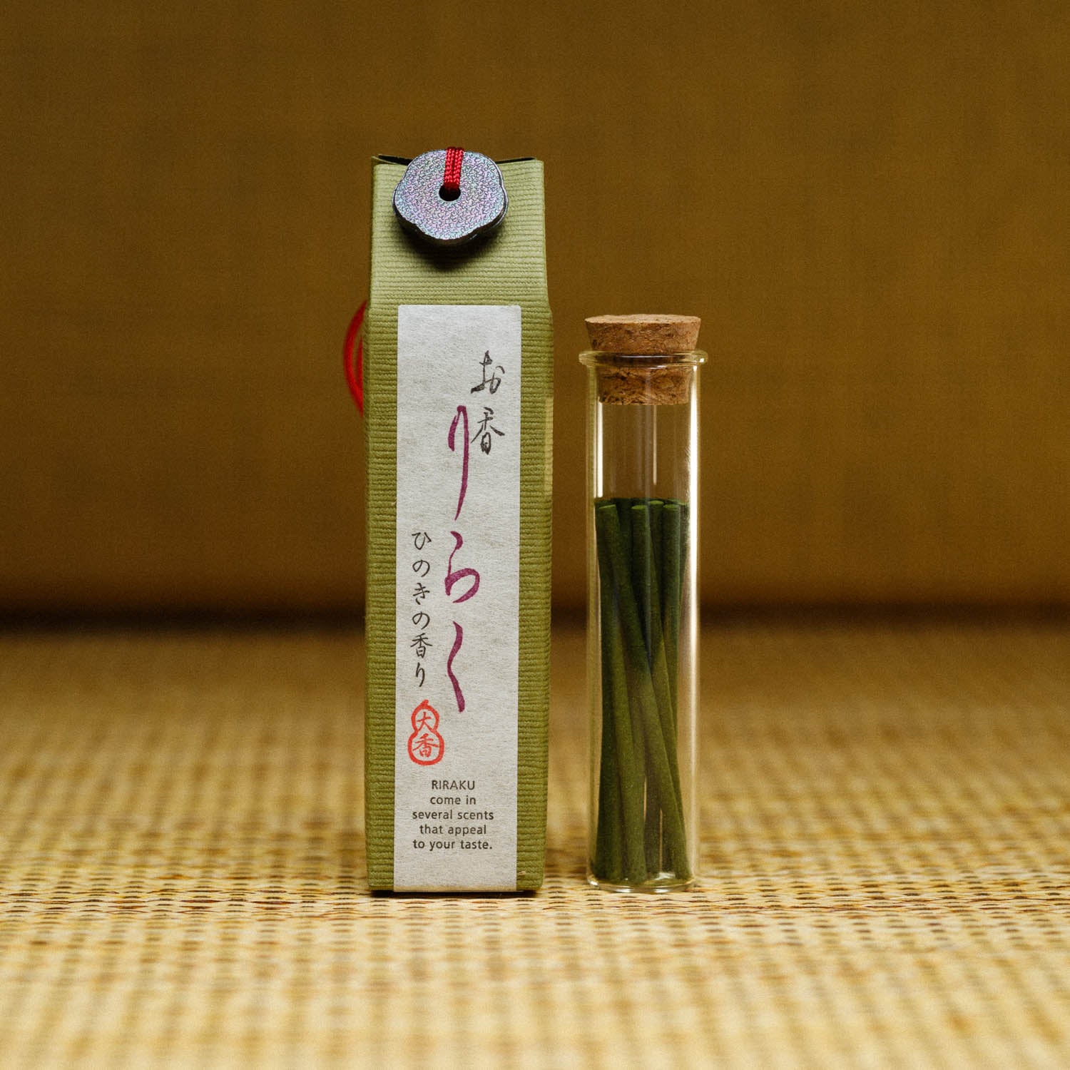 A small green box with Japanese characters labeled Riraku Hinoki Incense from Japan stands upright beside a sleek glass vial of thin green sticks. Decorative string and a circular seal adorn the box, set on a textured, woven background that evokes traditional Japanese fragrances.