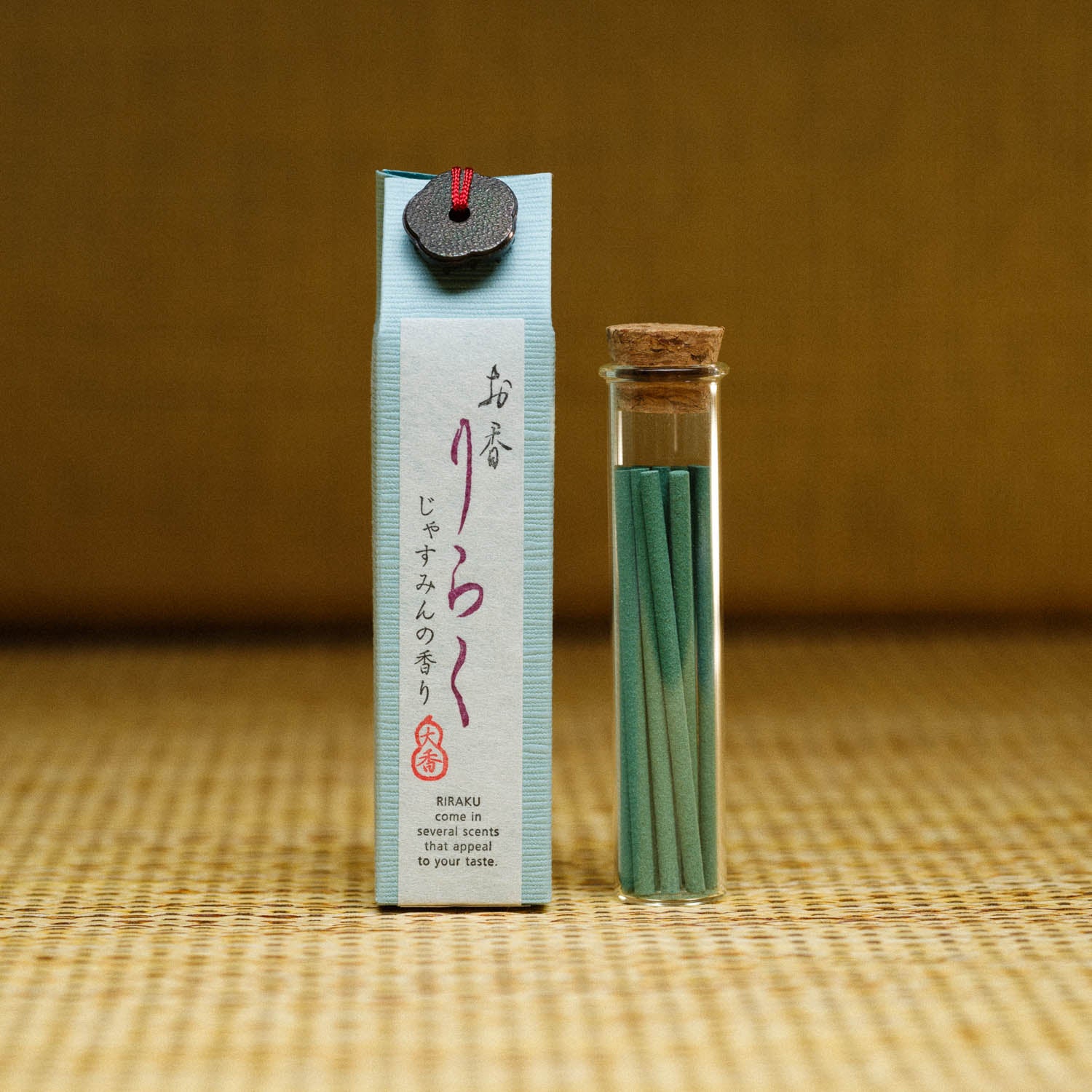 The Riraku Jasmine Incense from Japan features an elegantly designed small blue package highlighting Japanese fragrances, next to a glass vial with slender green incense sticks sealed with a cork. The background displays a textured tan surface and a muted dark green wall.