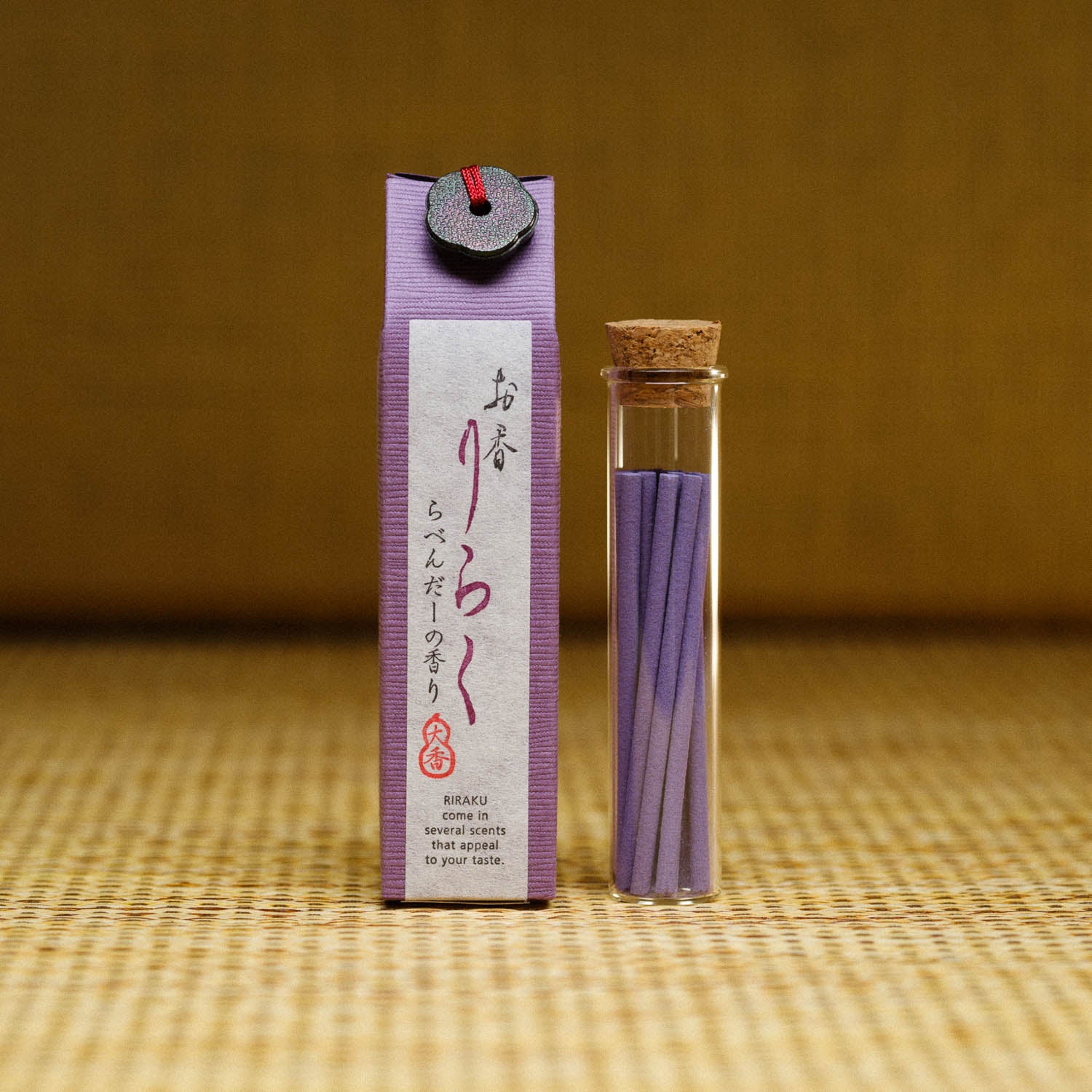 The Riraku Lavender Incense from Japan features thin, purple sticks in a small glass bottle with a cork. A purple rectangular box, decorated with Japanese writing and an illustration of a seated figure, complements the elegant display on a textured, neutral background.
