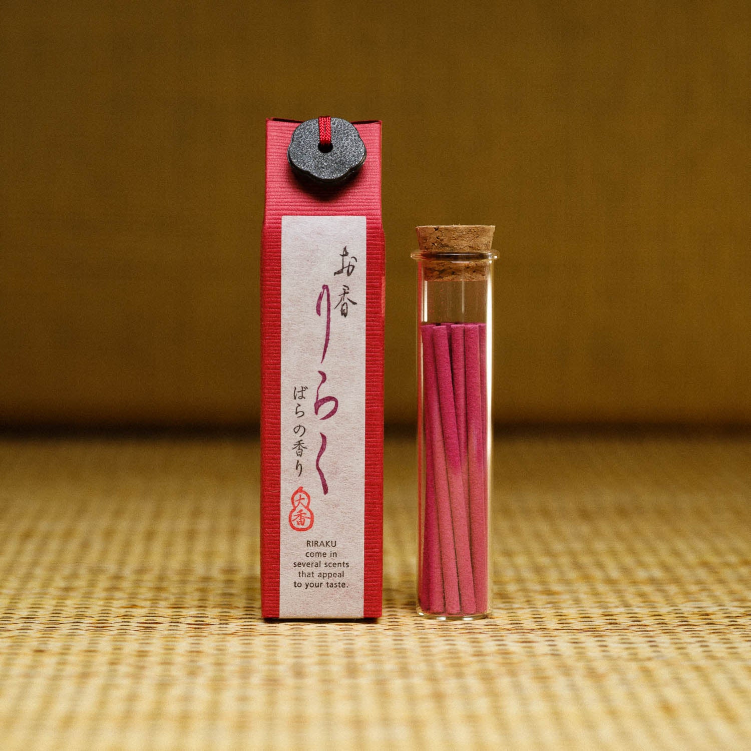 The Riraku Rose Incense from Japan features a red box adorned with Japanese text and Nippon Kodos emblem, accompanied by a glass vial of pink incense sticks sealed with a cork stopper, all displayed on a woven mat to highlight traditional Japanese fragrance packaging.