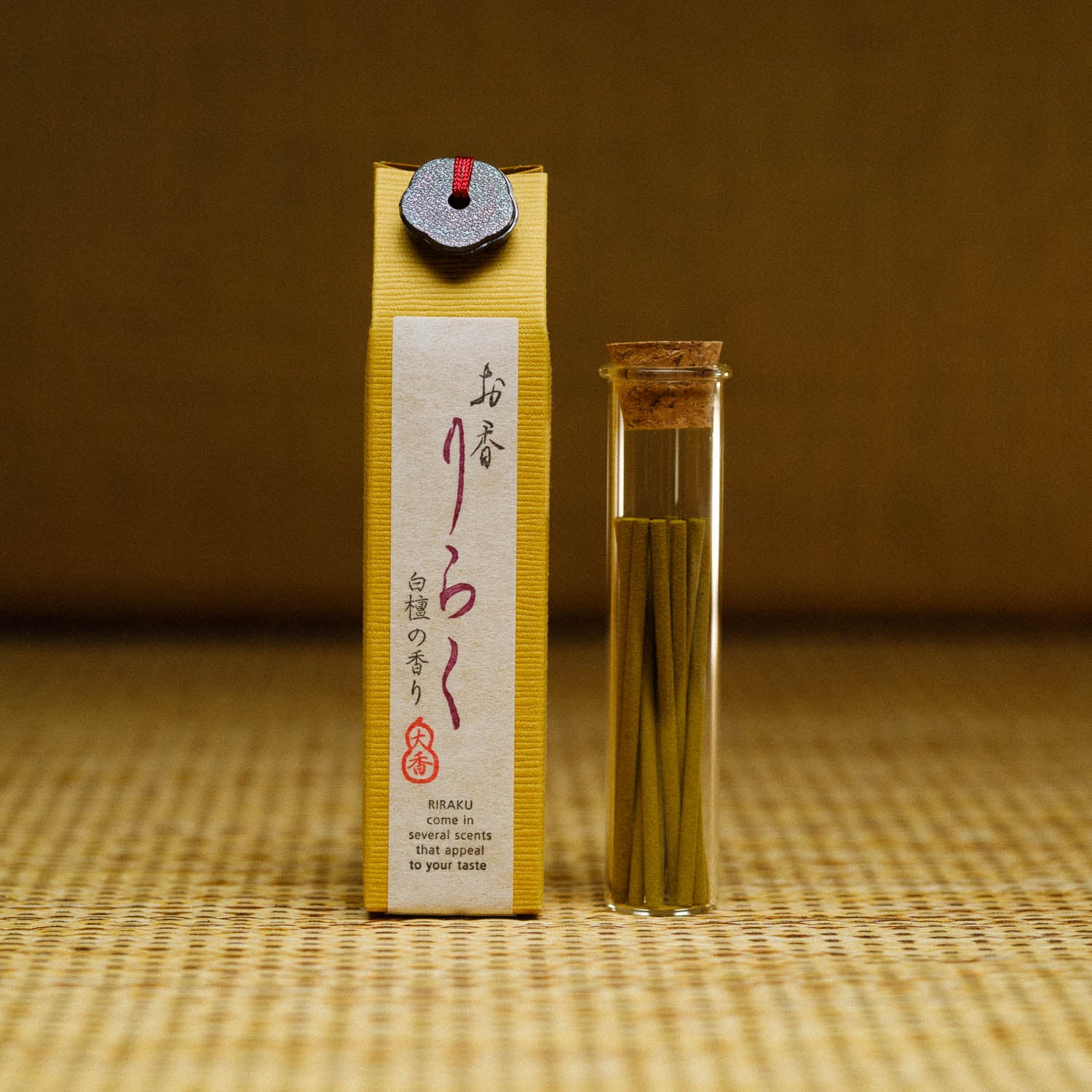 A package of Riraku Sandalwood Incense from Japan stands upright beside a glass tube with a cork stopper, containing thin sticks of incense. The packaging features Japanese text on a textured surface with traditional Nippon Kodo design against a woven background.