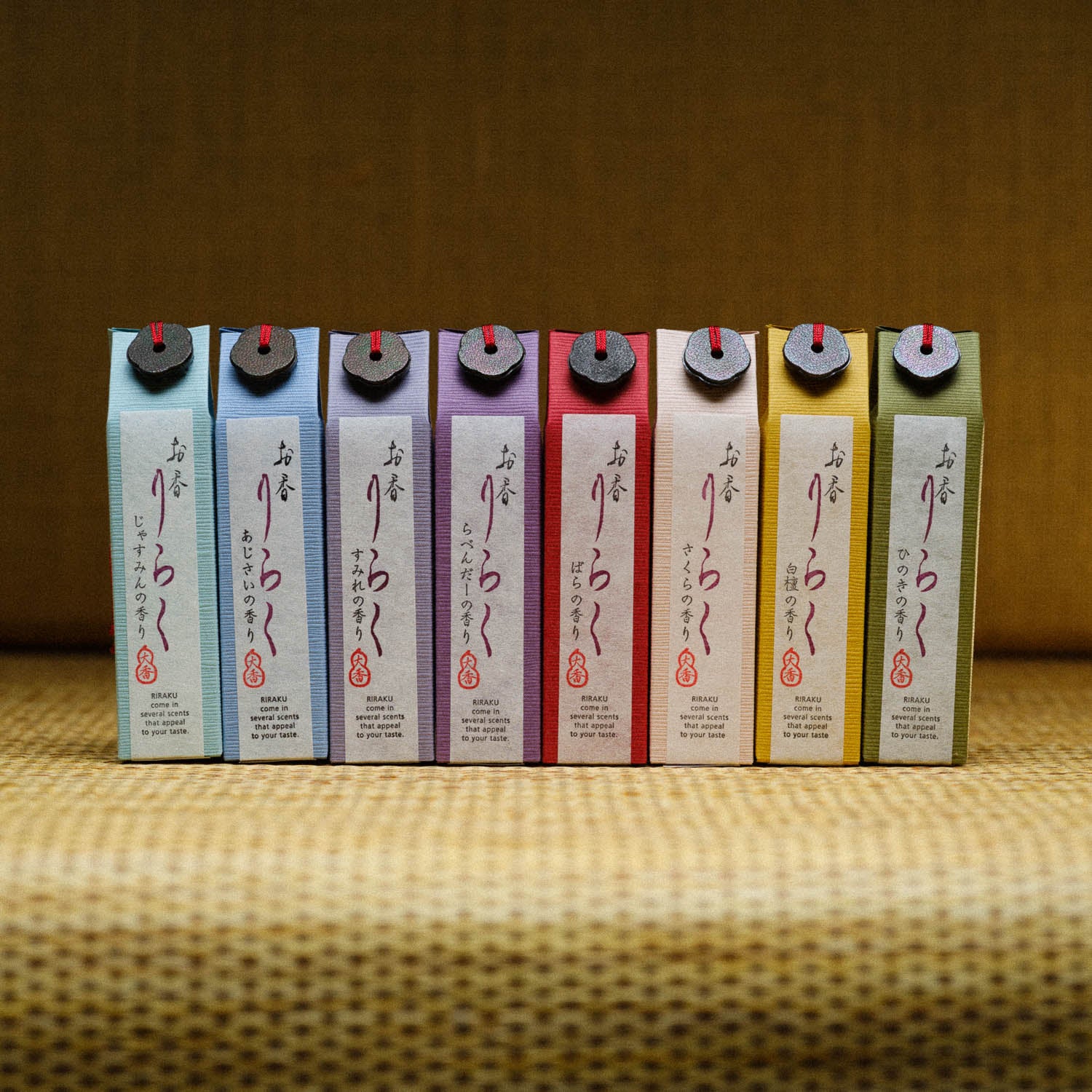 A row of Riraku Sandalwood Incense cartons from Japan, featuring colorful pastel tones, lines a textured brown background. Each carton, resembling traditional packaging, has vertical Japanese text and is adorned with a red ribbon or string on top.