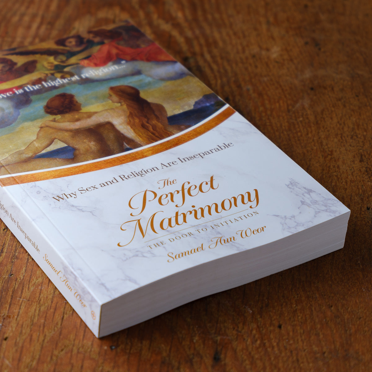 A close-up of Perfect Matrimony by Samael Aun Weor examines the link between sex and religion. The books marbled white cover, featuring people and a curved orange line evoking forbidden fruit, is gracefully displayed on a wooden surface.