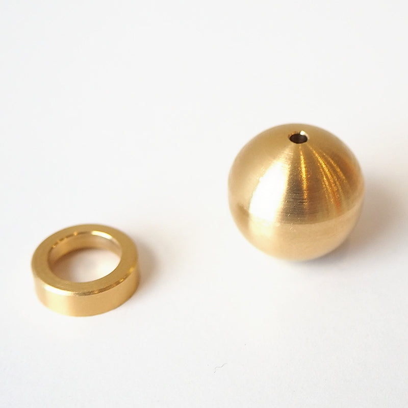 The Sphere Incense Holder, a smooth round brass ball with a small hole, sits elegantly on a white surface beside a brass ring. This Japanese-style incense holder features a shiny finish that highlights its solid metal craftsmanship, with the ball slightly larger than the ring.