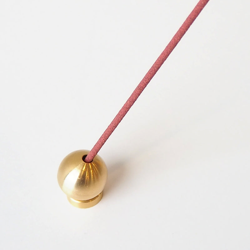 A close-up of the Sphere Incense Holder shows a shiny Japanese brass spherical design with a pink incense stick angled upwards on a white backdrop, blending tradition and elegance.
