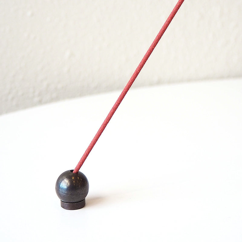 A red incense stick is elegantly angled in the Sphere Incense Holder, a small round brass piece, on a smooth white surface against a light gray background.