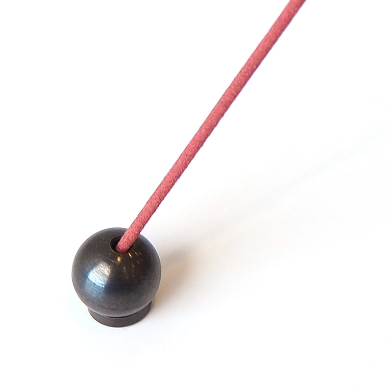 A red incense stick, set diagonally in a Sphere Incense Holder, creates a striking contrast against the plain white background.