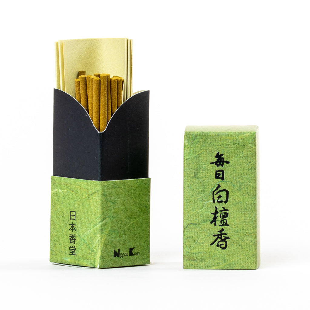 The Mainichi Byakudan Premium Sandalwood Japanese Incense comes in a green box with black Japanese text. Inside, brown sandalwood sticks are wrapped in gold paper, ideal for meditation. The textured pattern of the box adds an elegant touch.